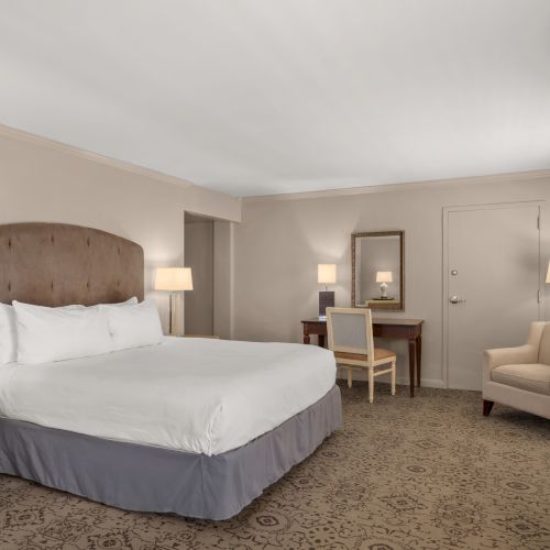 A hotel room with a king-size bed, a nightstand with lamps, a small desk with a mirror, and an armchair in a cozy setting.