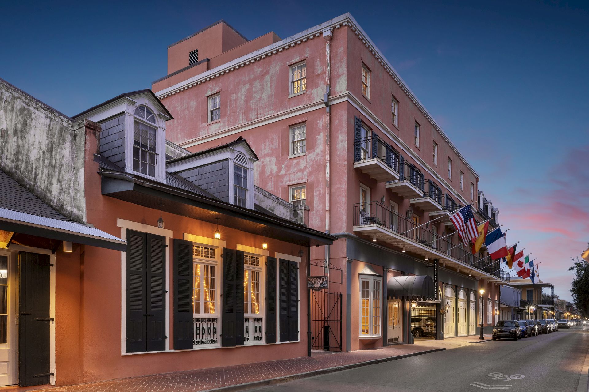 A historic building with a pink facade and balconies, adorned with international flags, lines a street at dusk.