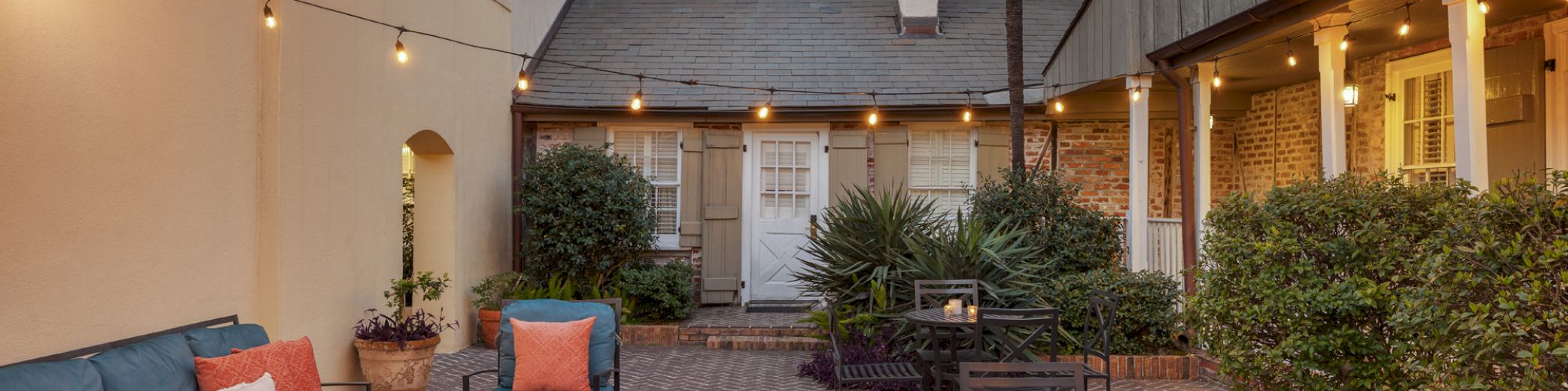 A cozy outdoor courtyard with string lights, comfortable seating, and tables, surrounded by plants and warm-toned buildings.