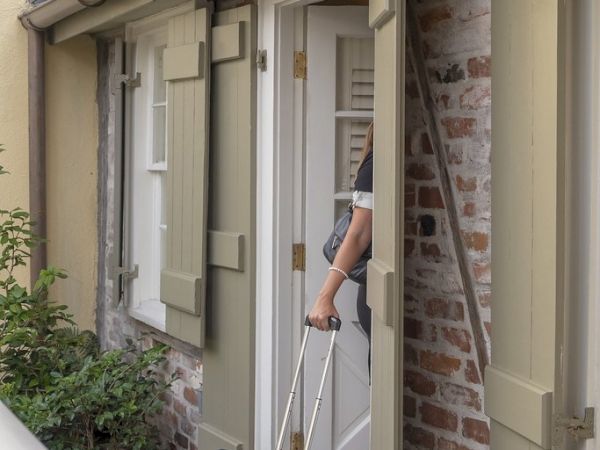 A person enters a brick building through a door, pulling a blue suitcase, with potted plants beside the path.