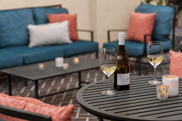 A cozy patio setup with a table, wine bottle, two glasses, candles, and colorful cushions on blue sofas.