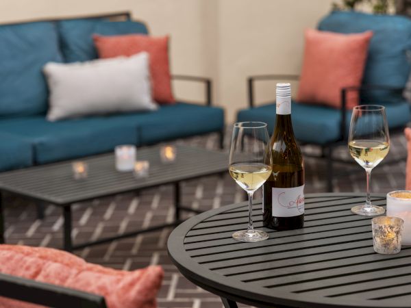 A cozy patio setup with a table, wine bottle, two glasses, candles, and colorful cushions on blue sofas.
