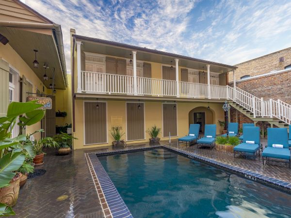 A charming courtyard features a pool, blue lounge chairs, and a two-story building with a balcony under a partly cloudy sky.