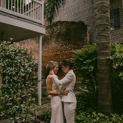 Two people in wedding attire embrace in a garden setting with brick and greenery in the background.