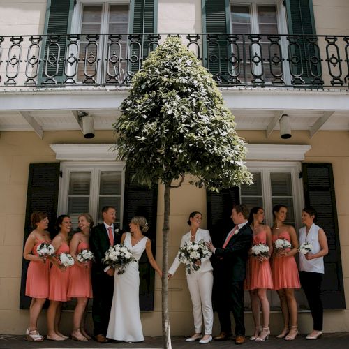 A group of people dressed formally, posing outside a building with a balcony and shuttered windows, some holding flowers.