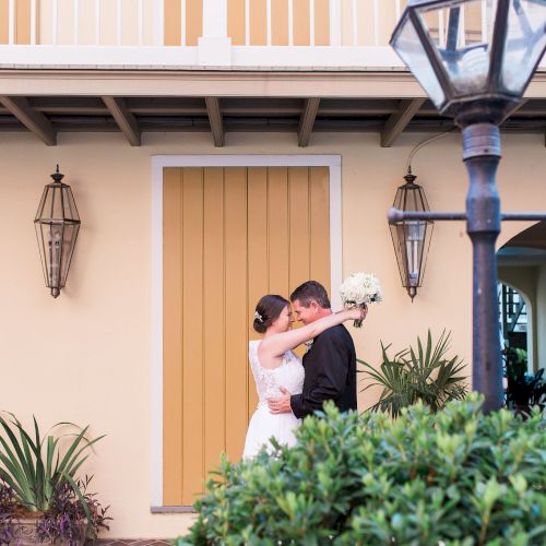 A couple embracing in front of a building with outdoor lights and plants, near a lamp post, under a balcony.