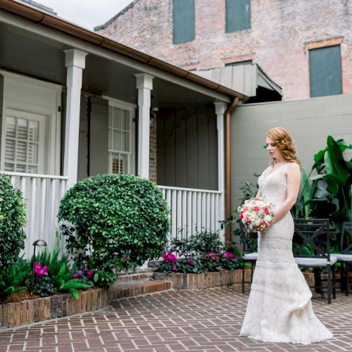 A bride in a white dress holds a bouquet, standing on a brick patio with greenery and a building in the background.