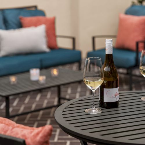 Patio setting with a black table, wine bottle, glasses, candles, and cushioned chairs in the background, creating a cozy atmosphere.