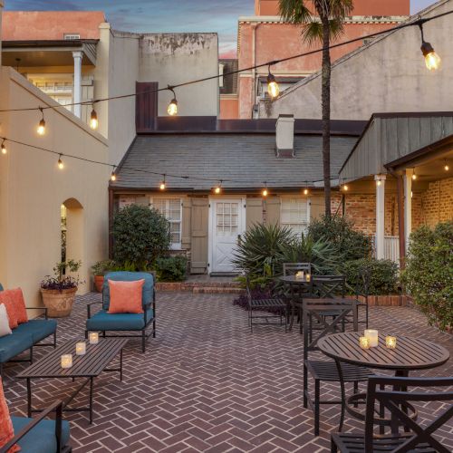 Outdoor patio with string lights, brick flooring, sofas, armchairs, tables, and candles; surrounded by buildings and plants; cozy ambiance.