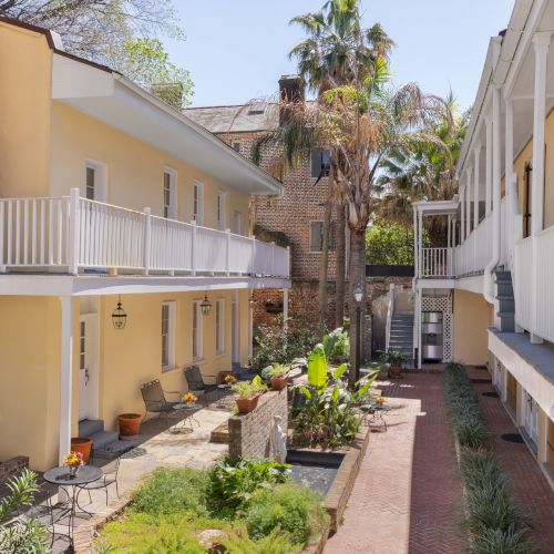 A charming courtyard with yellow buildings, white balconies, plants, and palm trees under a clear blue sky.
