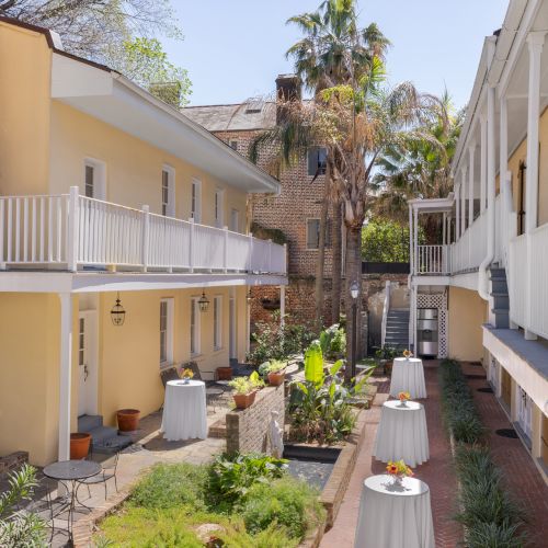 The image shows a courtyard between two yellow buildings with balconies, surrounded by palm trees and plants, and tables set with flowers.