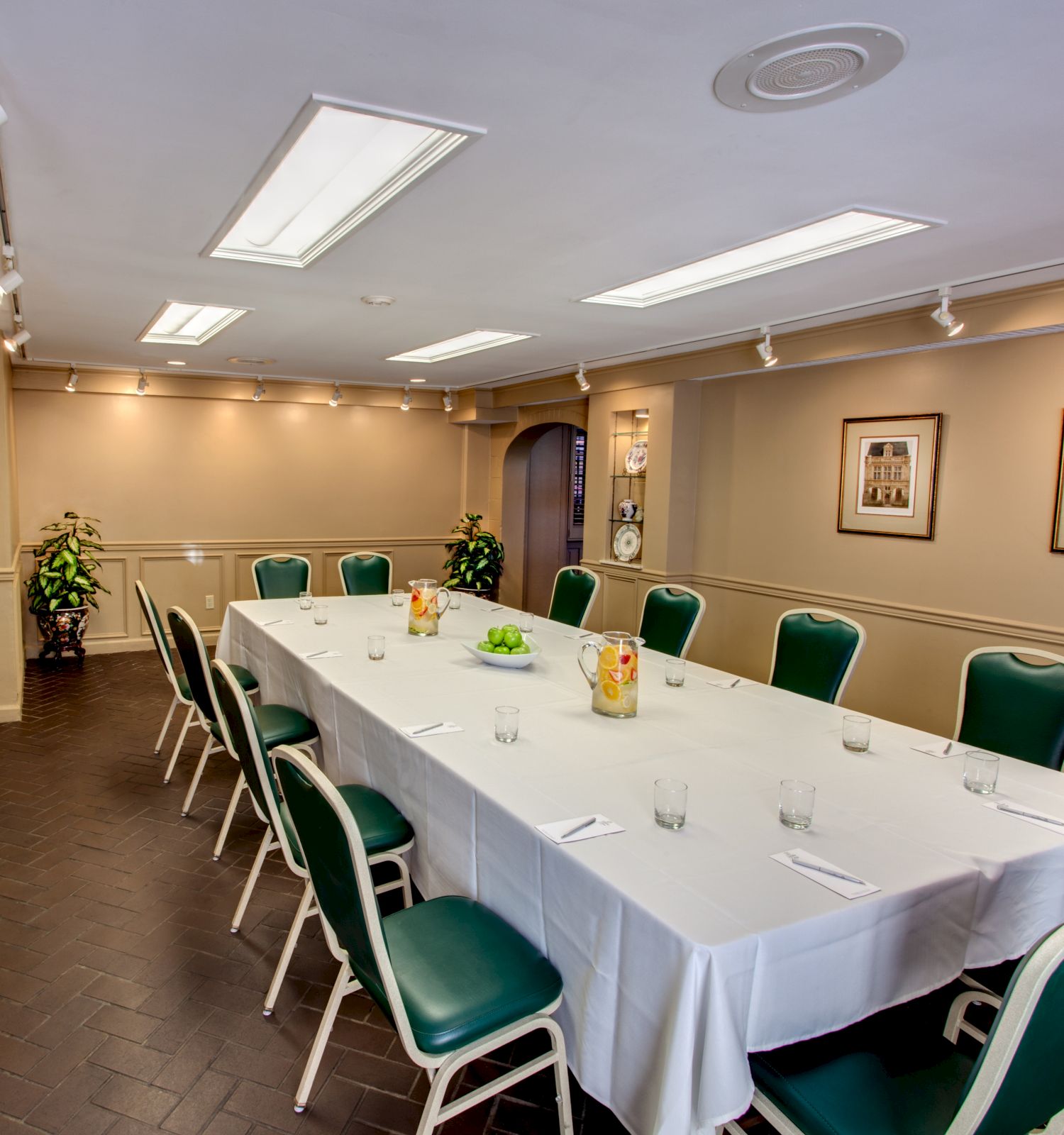 The image shows a conference room with a long table, green chairs, pitchers of drinks, and framed artwork on the walls.