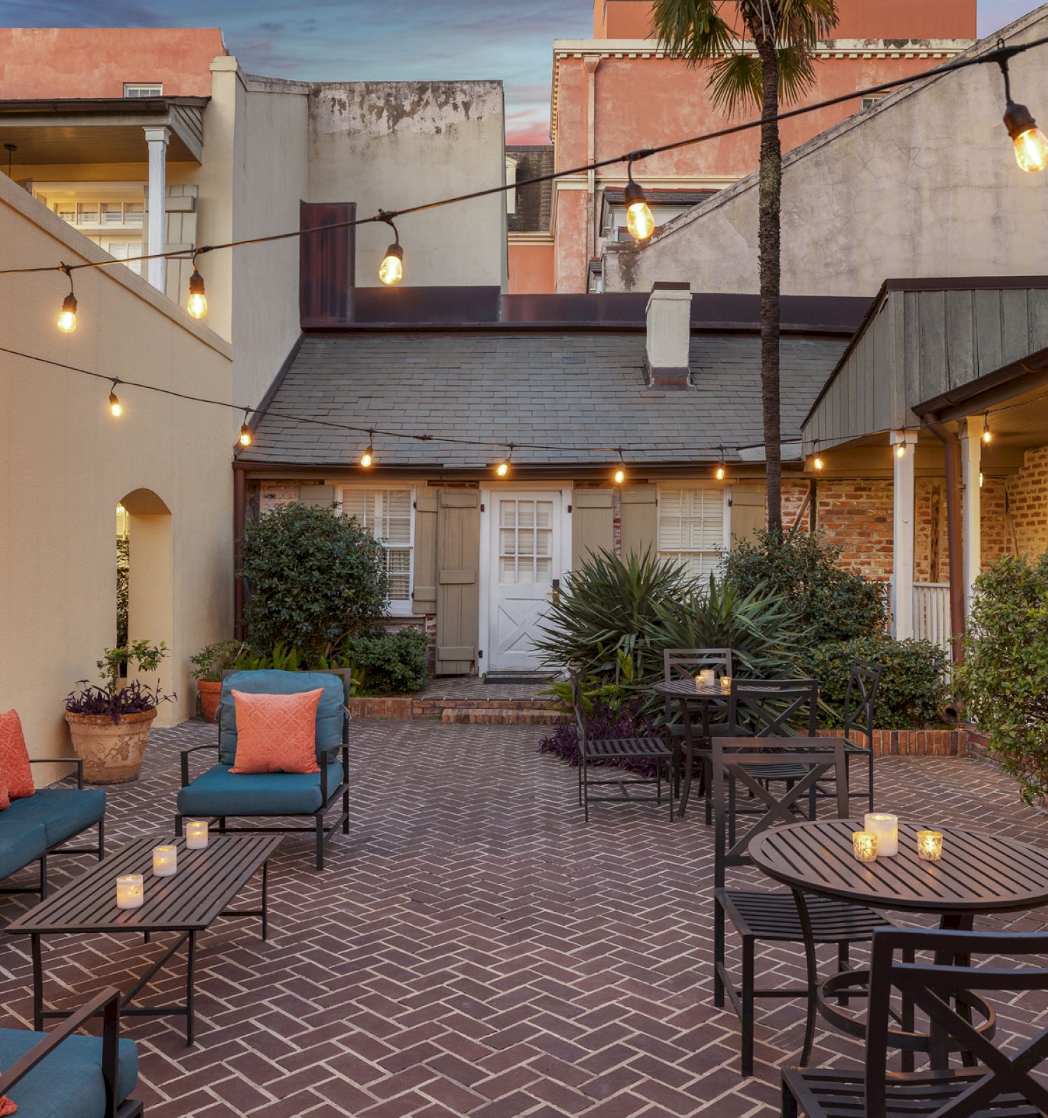 A cozy patio with string lights, outdoor seating, potted plants, and candles on tables, surrounded by buildings.