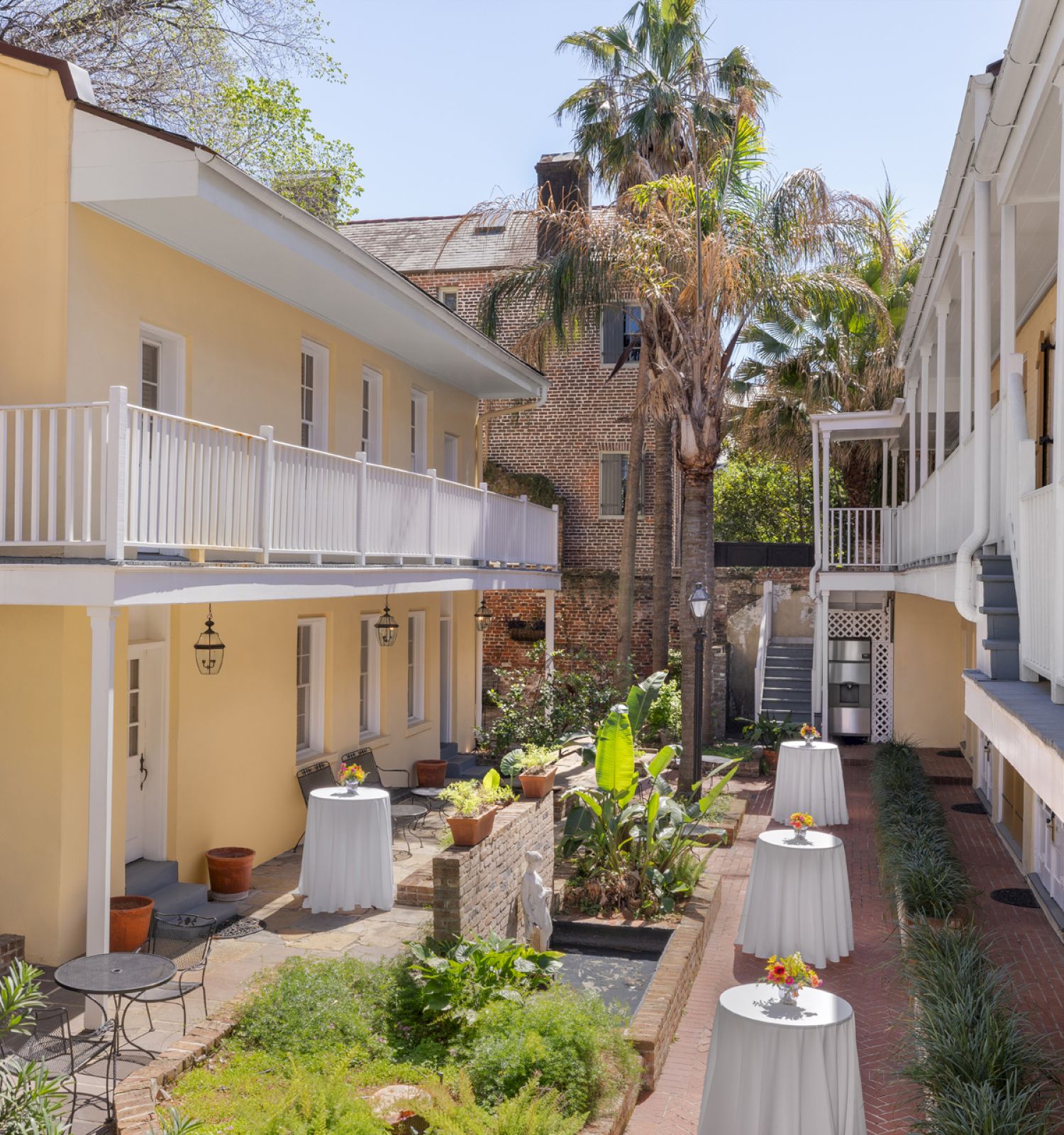 A charming outdoor courtyard with yellow buildings, white railings, potted plants, and tables with white tablecloths and floral centerpieces.