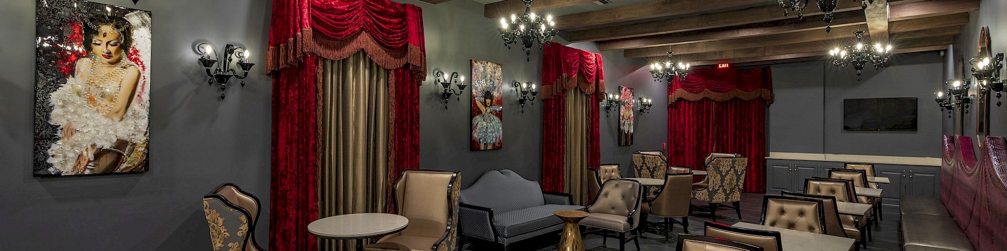 A stylish lounge with elegant chandeliers, red curtains, decorative art, plush seating, and wooden ceiling beams.