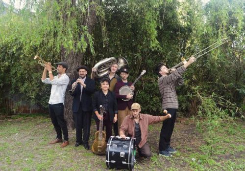 A group of seven people outdoors, holding various musical instruments, including a trumpet, tuba, and banjo, stand in front of a tree.