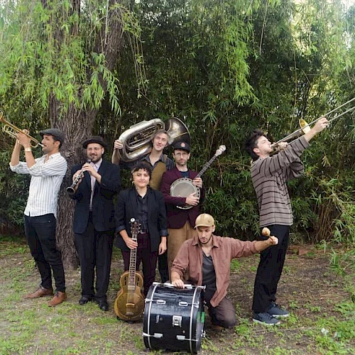 A group of seven people outdoors, holding various musical instruments, including a trumpet, tuba, and banjo, stand in front of a tree.