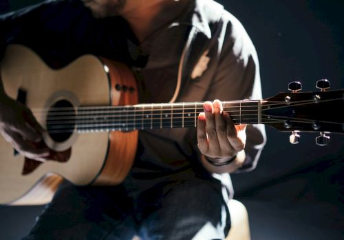 A person is playing an acoustic guitar, focusing on the fingers placed on the fretboard in a dimly lit setting.
