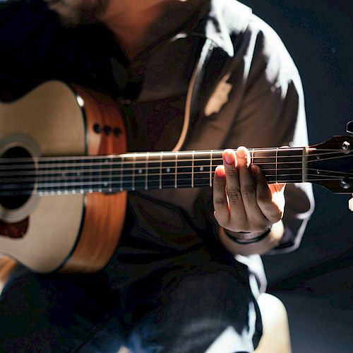 A person is playing an acoustic guitar, with focus on their hands and the guitar's strings and body, in a dimly lit setting.