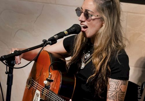 A person wearing sunglasses is singing and playing an acoustic guitar with a microphone in front of them.