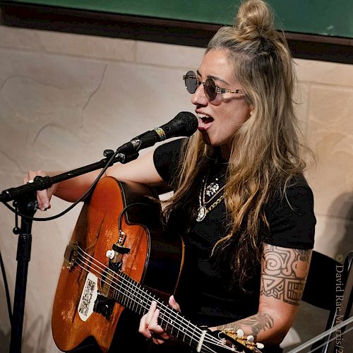 A person wearing sunglasses is singing and playing an acoustic guitar with a microphone in front of them.