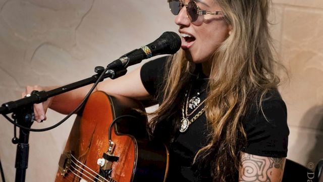 A person wearing sunglasses is singing and playing an acoustic guitar with a microphone in front of them.