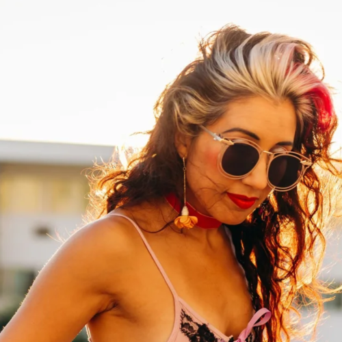 A person with sunglasses and colorful hair, wearing earrings and a choker, poses outside with a sunny backdrop.