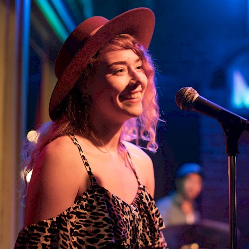 A person in a hat smiling by a microphone on stage, with colorful lights creating a lively atmosphere.