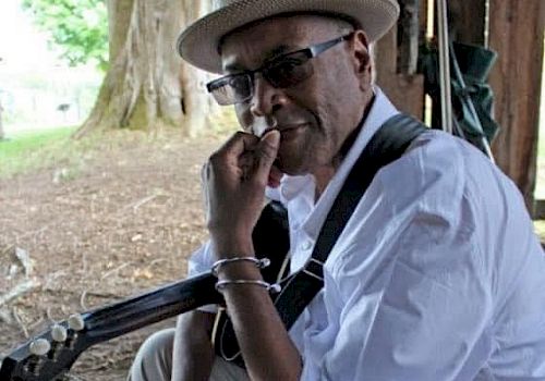 A person wearing a hat and glasses sits under a tree, holding a guitar and resting their chin on their hand, with a thoughtful expression.