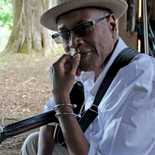 A person wearing a hat and glasses sits under a tree, holding a guitar and resting their chin on their hand, with a thoughtful expression.