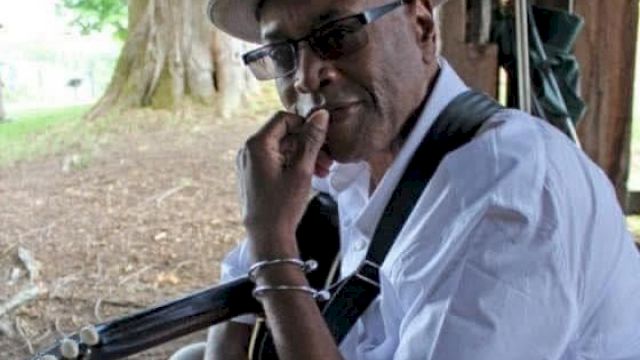 A person wearing a hat and glasses sits under a tree, holding a guitar and resting their chin on their hand, with a thoughtful expression.