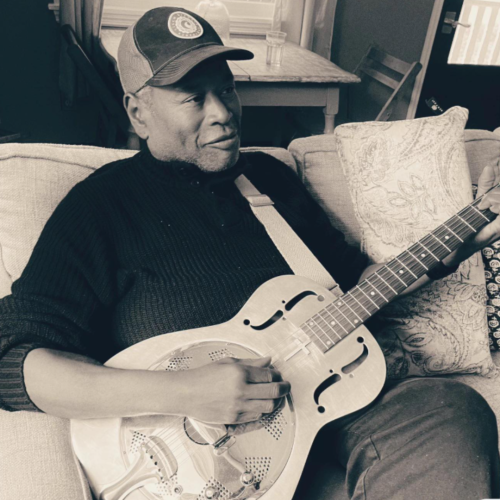 A person wearing a cap is sitting on a couch, playing a resonator guitar.