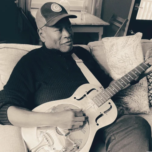 A person wearing a cap is sitting on a couch, playing a resonator guitar.