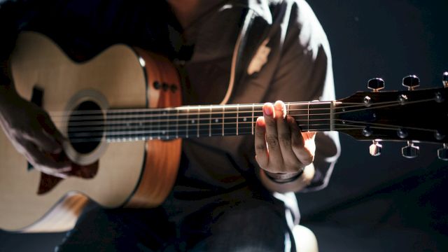 A person is playing an acoustic guitar, focusing on the fretboard with a dimly lit background.