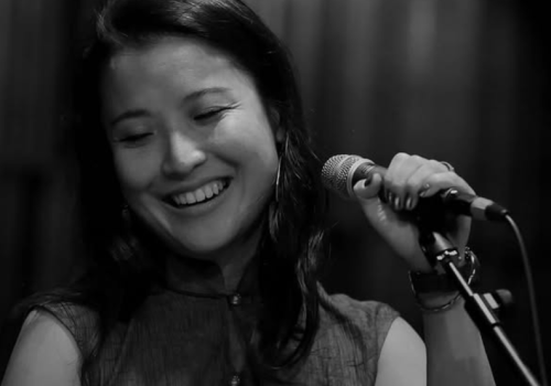 A person smiling while holding a microphone, seemingly engaged in a musical or vocal performance, captured in black and white.