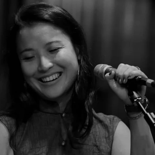 A person smiling while holding a microphone, seemingly engaged in a musical or vocal performance, captured in black and white.