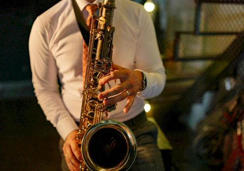 A person is playing a saxophone, wearing a white shirt and tie, in a dimly lit setting with stairs in the background.