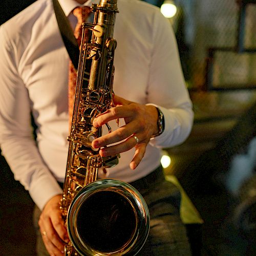 A person is playing a saxophone, wearing a white shirt and tie, in a dimly lit setting with stairs in the background.