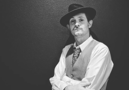A person in vintage attire, wearing a suit, tie, and fedora, poses against a wall in a black and white photo.