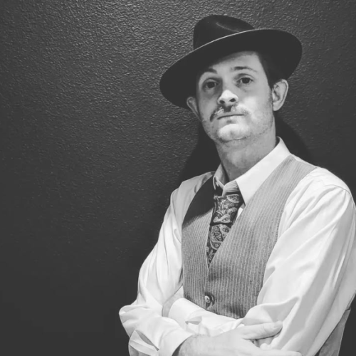 A person in vintage attire, wearing a suit, tie, and fedora, poses against a wall in a black and white photo.