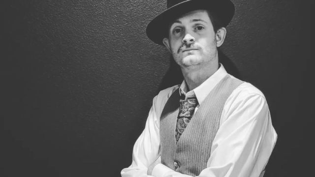 A person in vintage attire, wearing a suit, tie, and fedora, poses against a wall in a black and white photo.