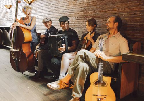 A group of five musicians with instruments, including a double bass, accordion, violin, and guitar, seated and standing in a cozy setting.