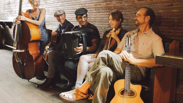 A group of five musicians with instruments, including a double bass, accordion, violin, and guitar, seated and standing in a cozy setting.