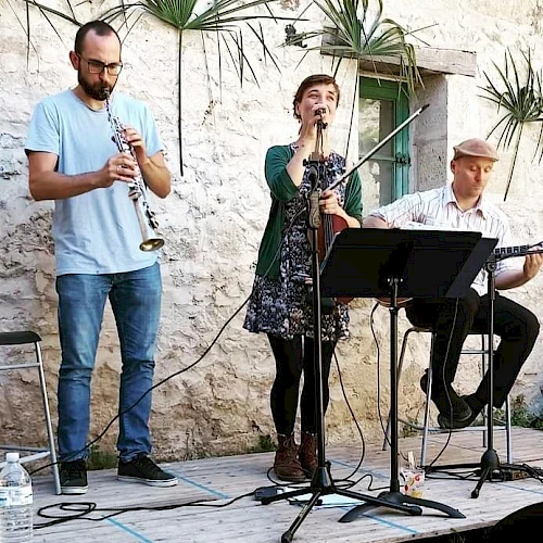 A trio of street musicians performs outside a rustic stone wall: a saxophonist in blue, a drummer-singer with a guitar, and a seated guitarist with sheet music, outdoors.