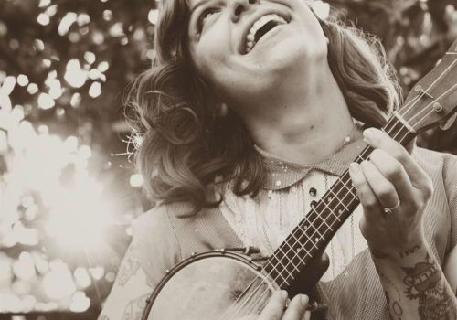 A smiling woman plays a small string instrument (a ukulele) outdoors, soaking in sunlight and joy.