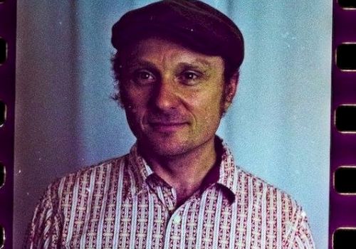 A man wearing a patterned button-up shirt and a dark hat, standing indoors with a blue backdrop, film-strip edges visible in the frame.