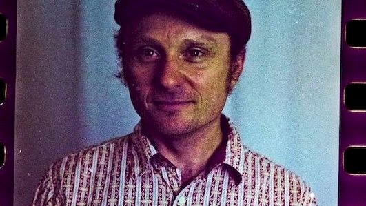 A man wearing a patterned button-up shirt and a dark hat, standing indoors with a blue backdrop, film-strip edges visible in the frame.
