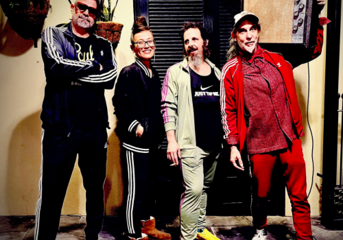 Four casually dressed friends posing together indoors, some with bright sneakers, smiling and standing in a row.