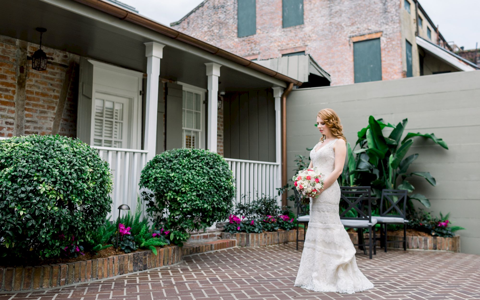 A bride in a white gown holds a bouquet, standing on a brick patio by a building, surrounded by greenery and flowers.
