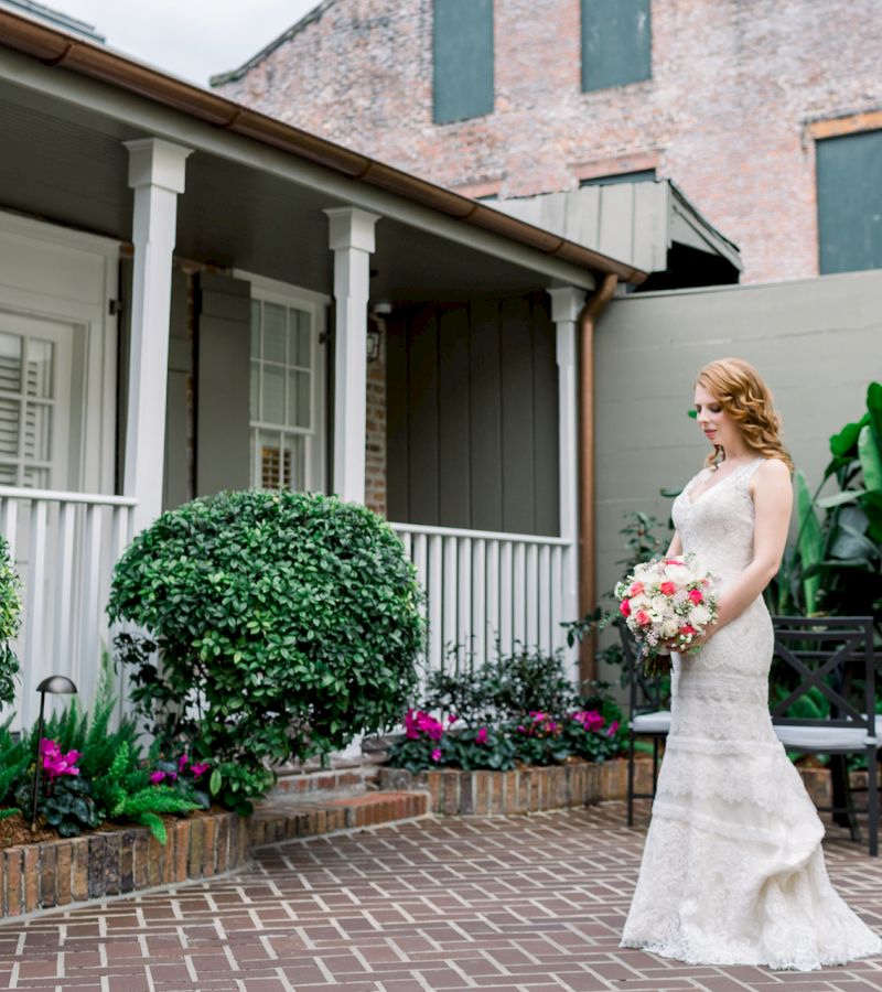 A woman in a wedding dress stands on a brick patio in front of a house with a porch.